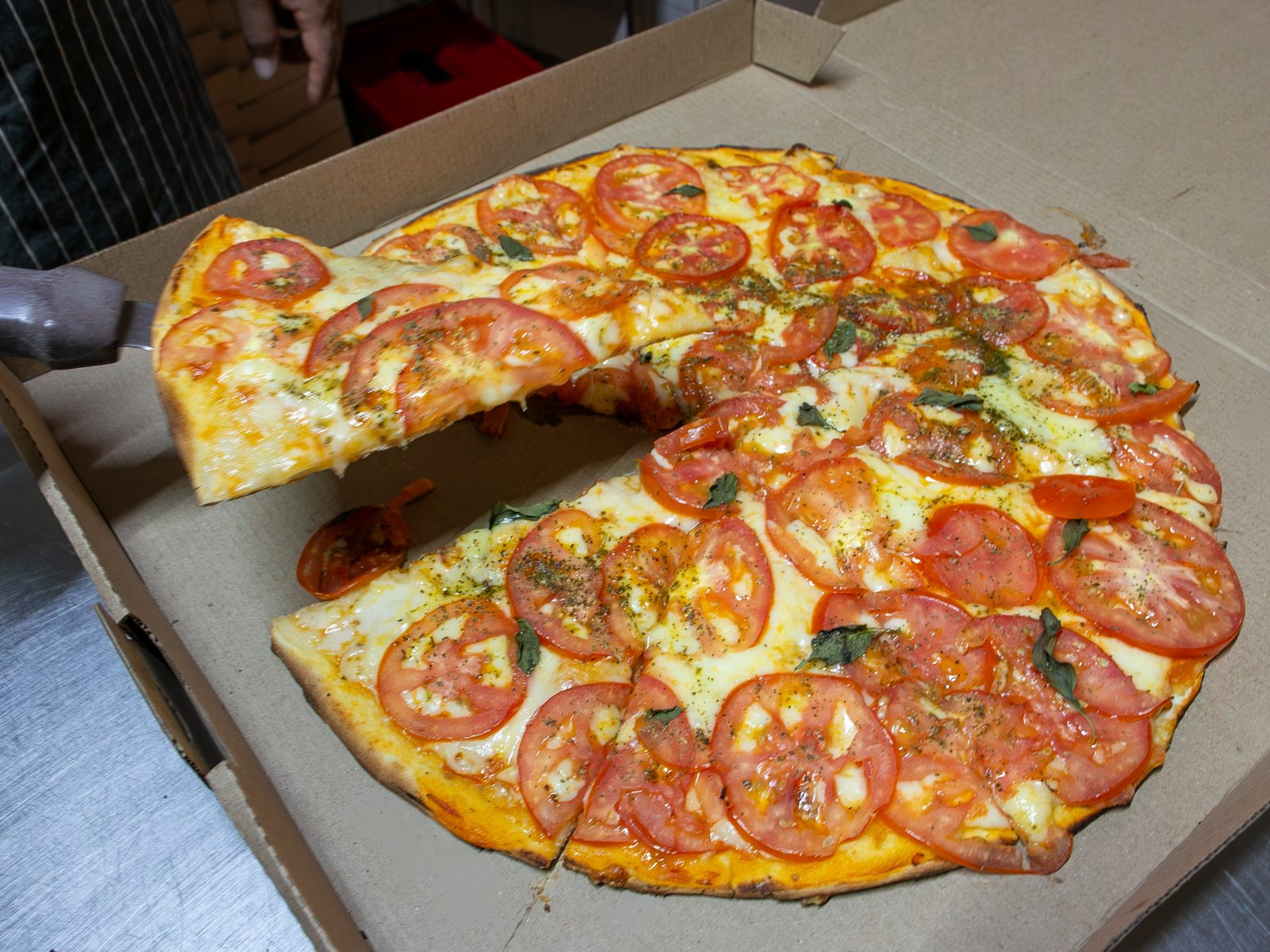 Pizza margarita con jitomate fresco, mozzarella y albahaca recién horneada en su caja para llevar, una rebanada levantándose